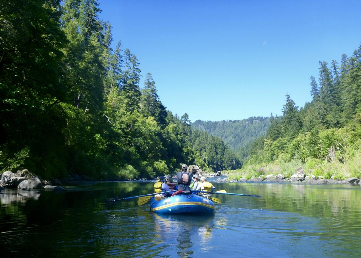 Five people on an inflatable raft float between forest covered shores.