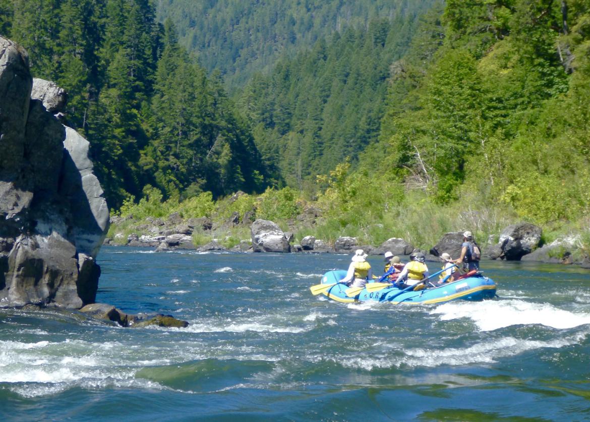 Six people on an inflatable raft paddle on a river flowing through a forest.
