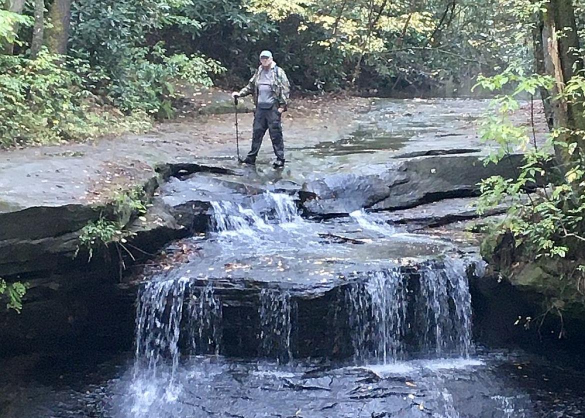 A hiker stepping on the top of the running stream while holding trekking poles.
