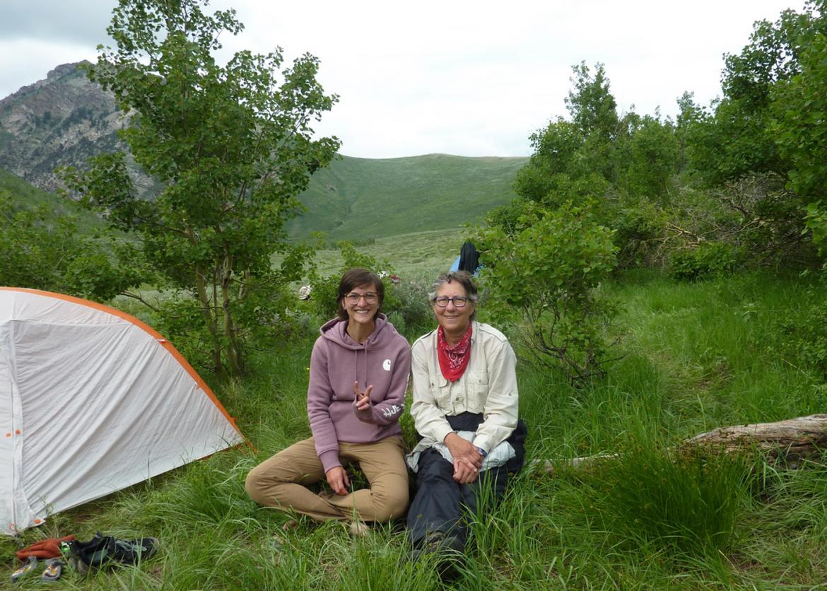 Two women sitting and smiling on the grass field next to their tent.