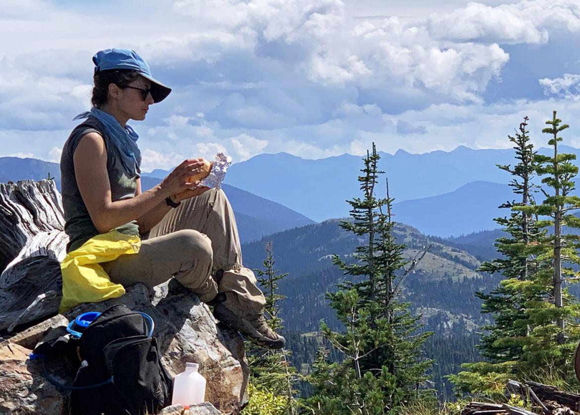 A hiker in sunglasses sitting at the edge of a cliff, holding a snack.