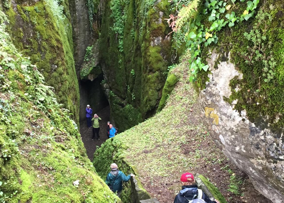 Hikers walk down a trail between close stone walls covered in moss.