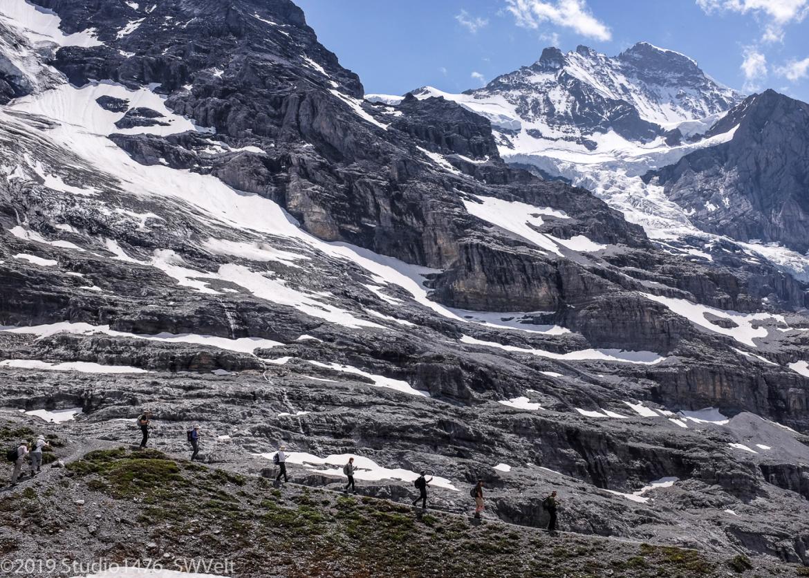 Hikers walk on a trail on rocky face of a snowy mountain.