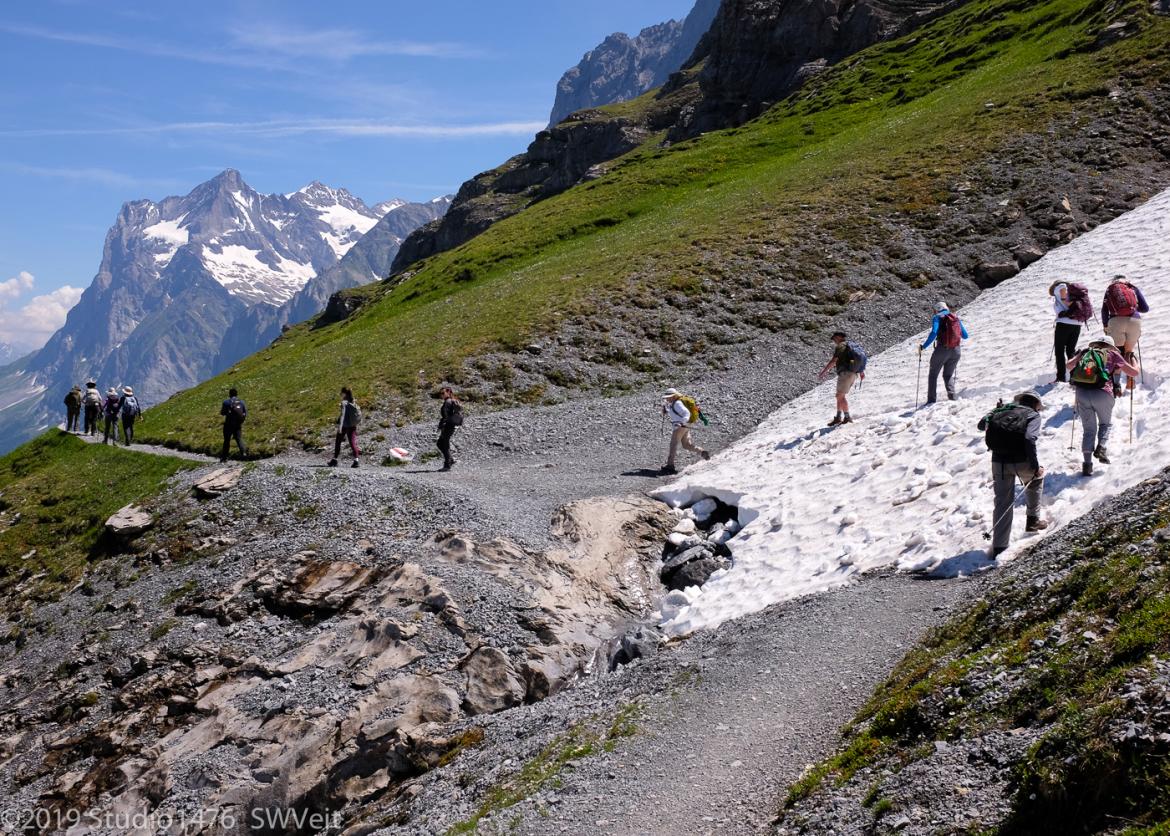 Hikers carefully pick their way across a slope which is alternatively covered in snowy, gravel, and grass.