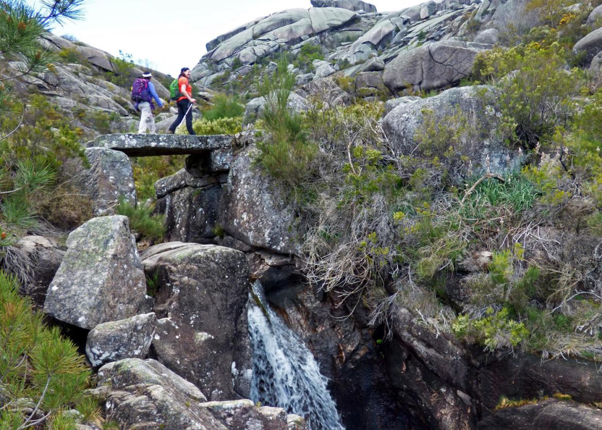 Two people walk over a flat stone serving as a bridge.  Below is a small waterfall, and surrounding them are rocky hillsides. Twiggy plants grow from the rocks.