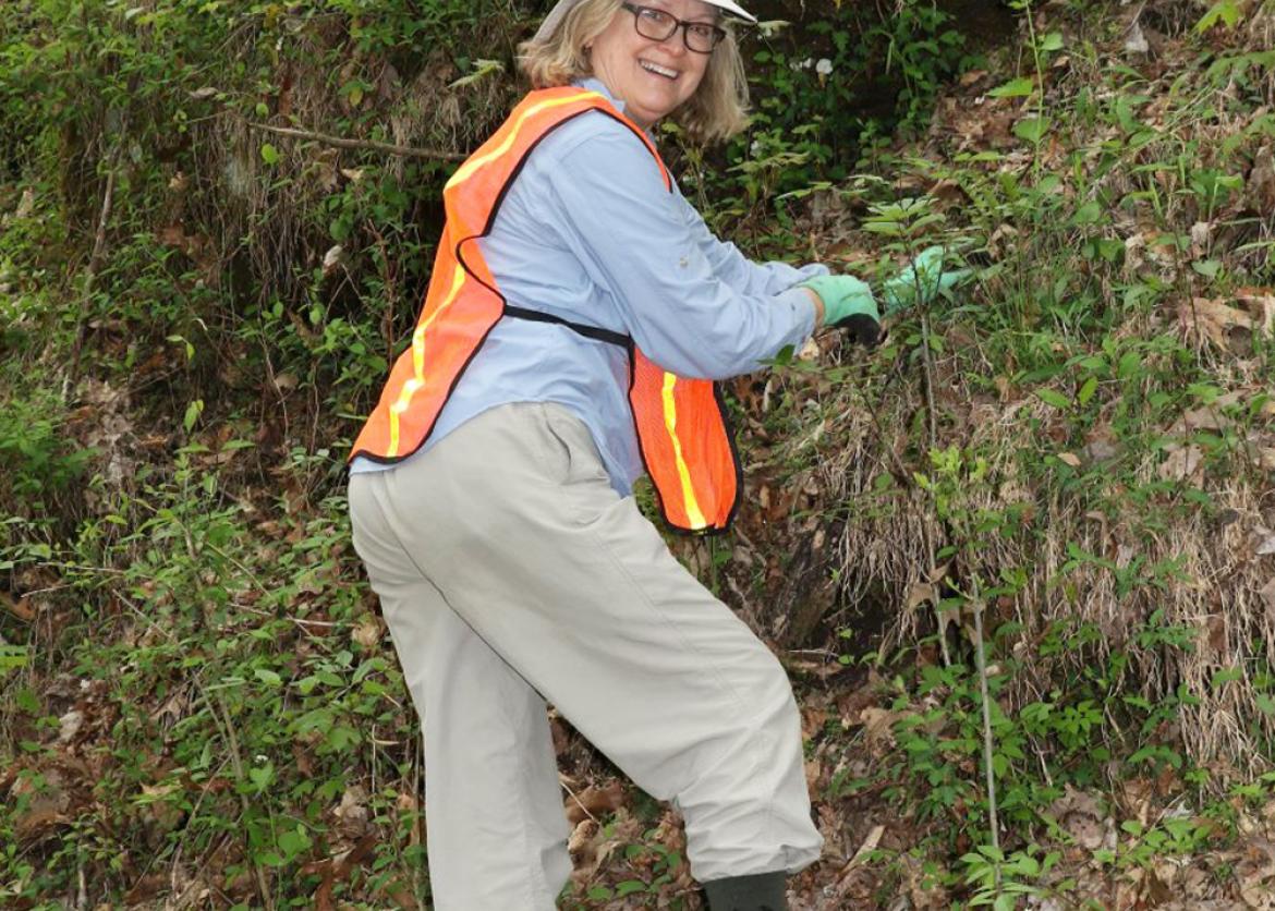 A woman in an orange vest and work gloves turns back and smiles.