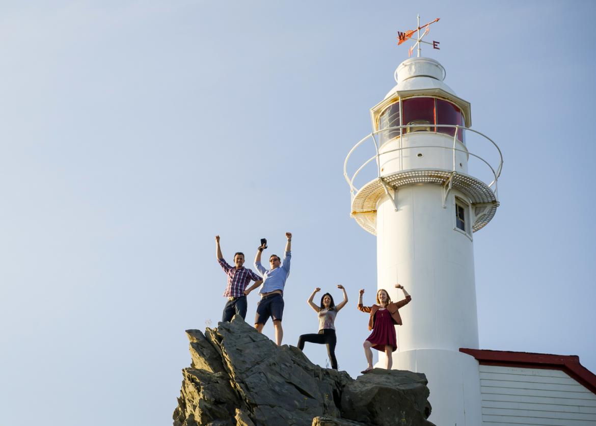 Four people on a rock, posing with their arms with the lighthouse behind them.