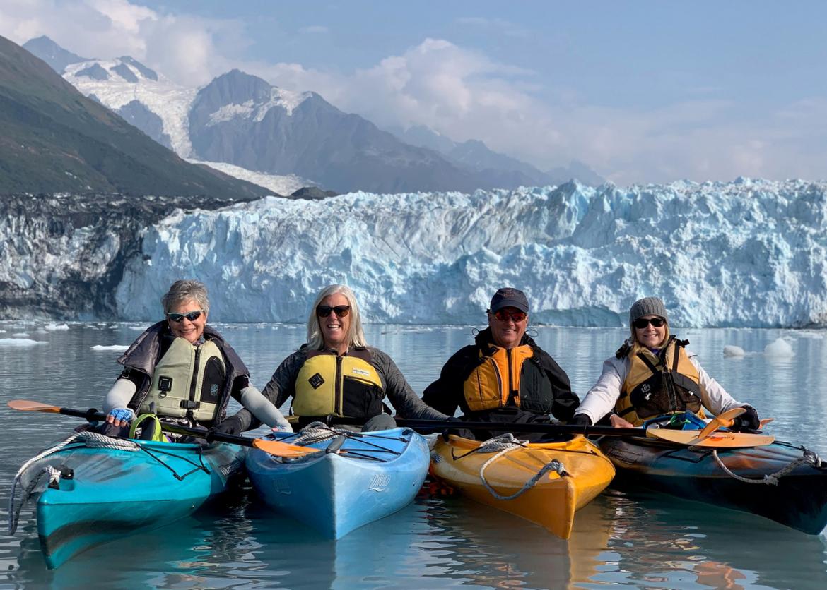 Four smiling kayakers in the water hold each other in place to float in a row. Behind them is a frozen outcropping and mountains.