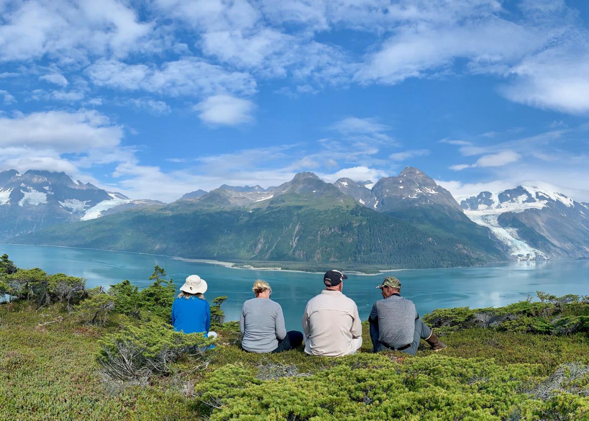 Four people sit next to each other overlooking bright water and forested mountainside.