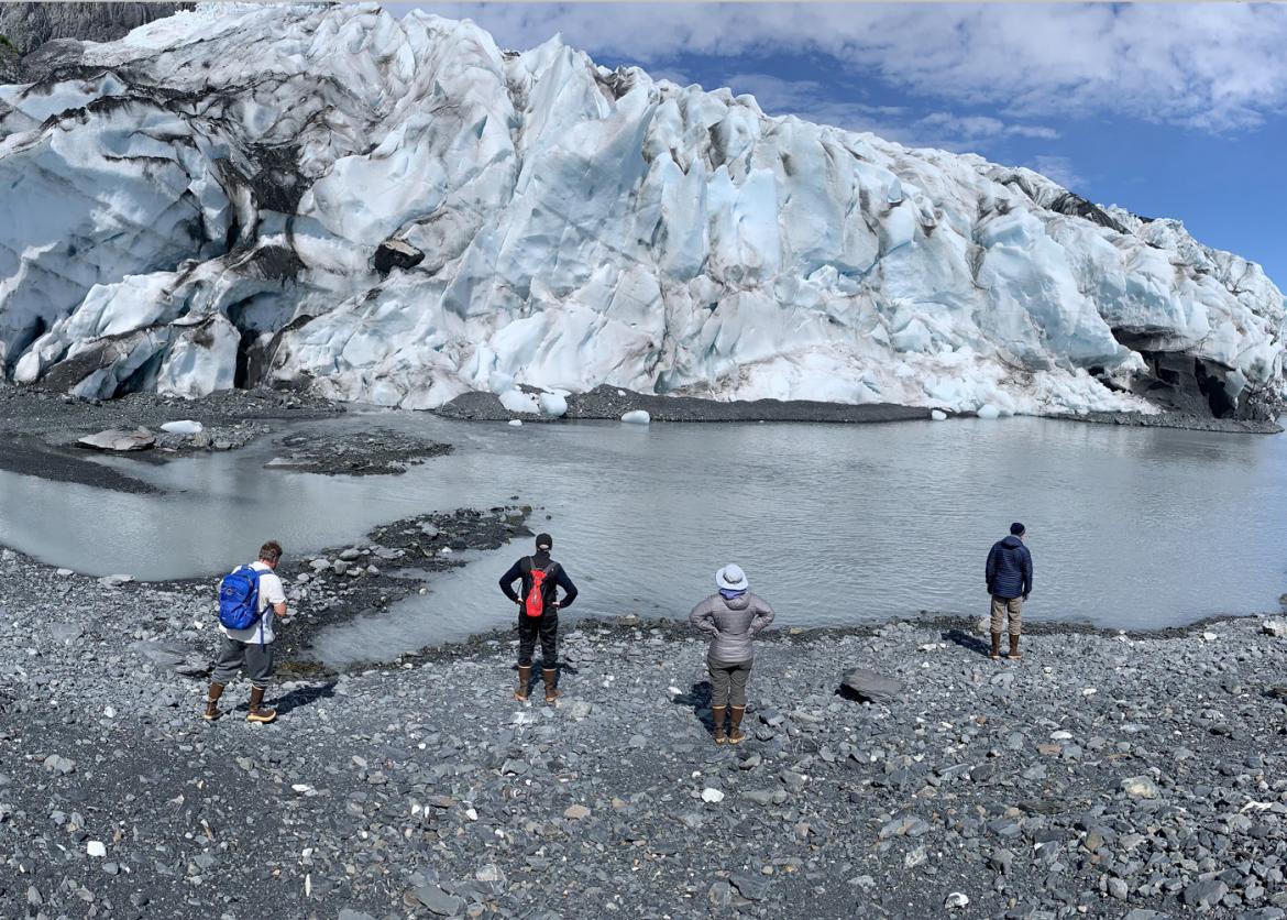 Four people standing on a rocky shore observe icy waters. On the other side of the water rises icy rocks.