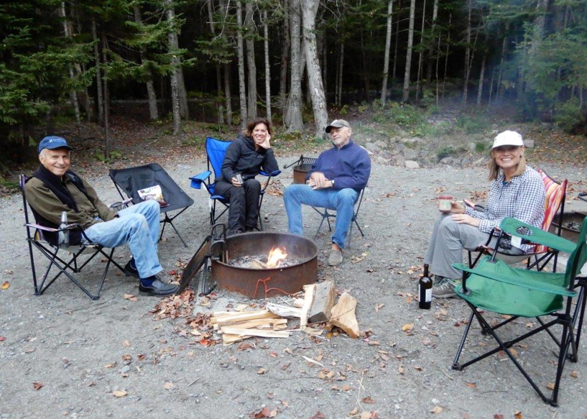 A group of people sitting and smiling, surrounding the fire outdoors