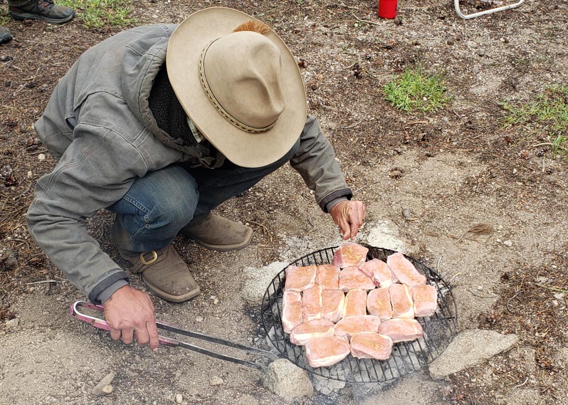 A man in a cowboy hat, squatting down, adding seasoning to the chicken on the barbeque grill