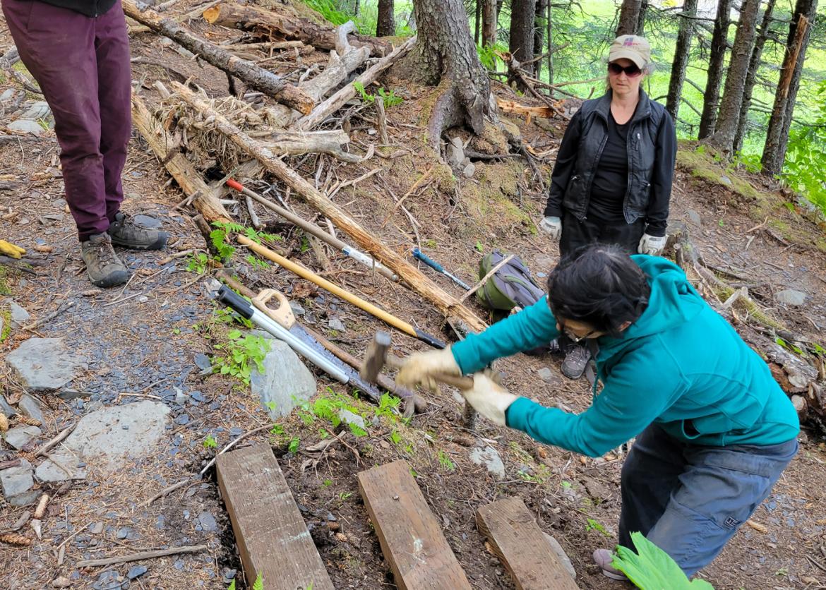 Two people watch a third person swing a hammer at wooden trail steps.