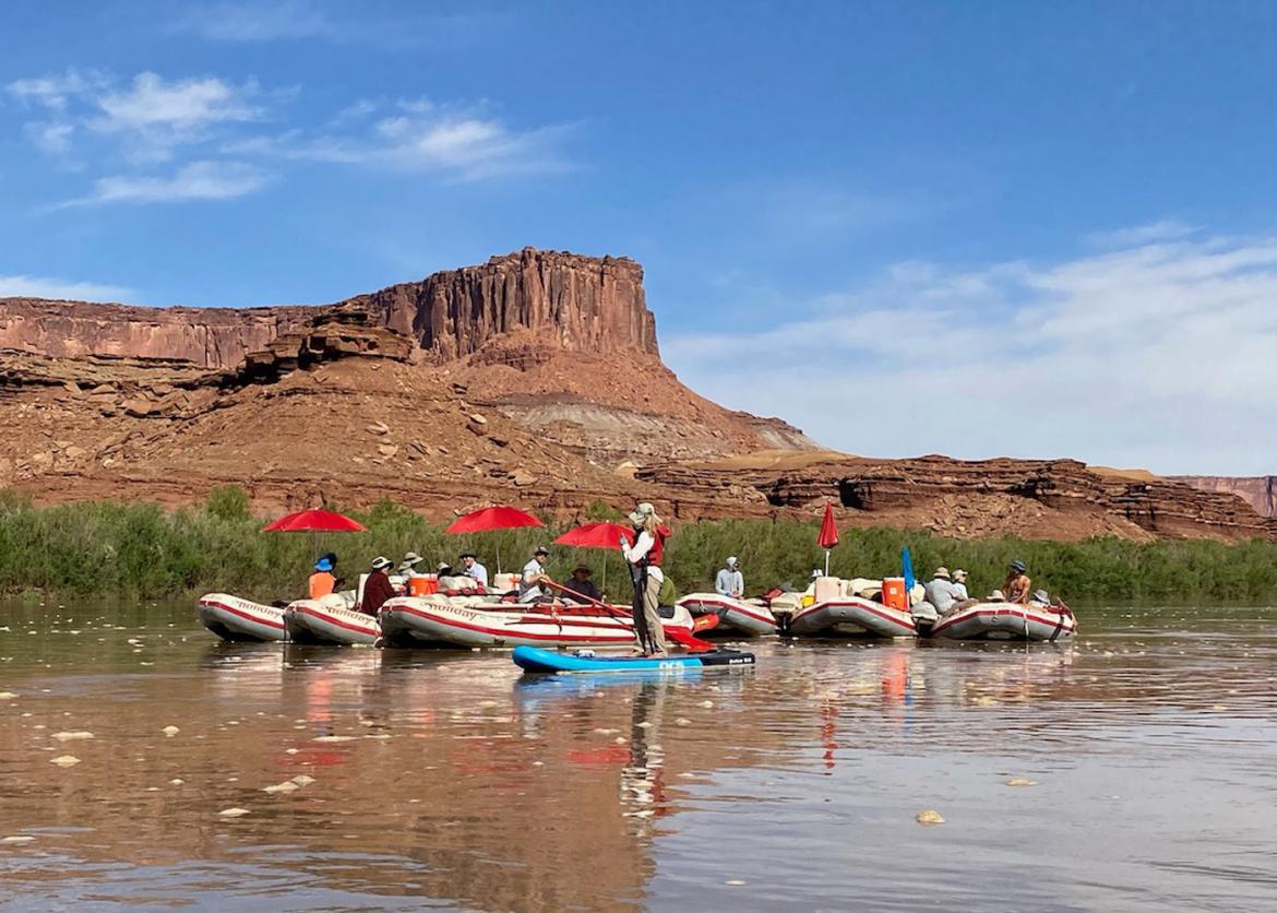 Six inflatable rafts, some shaded by umbrellas. A single woman stands on a paddleboard. Beyond the water's edge lies a canyon ridge.