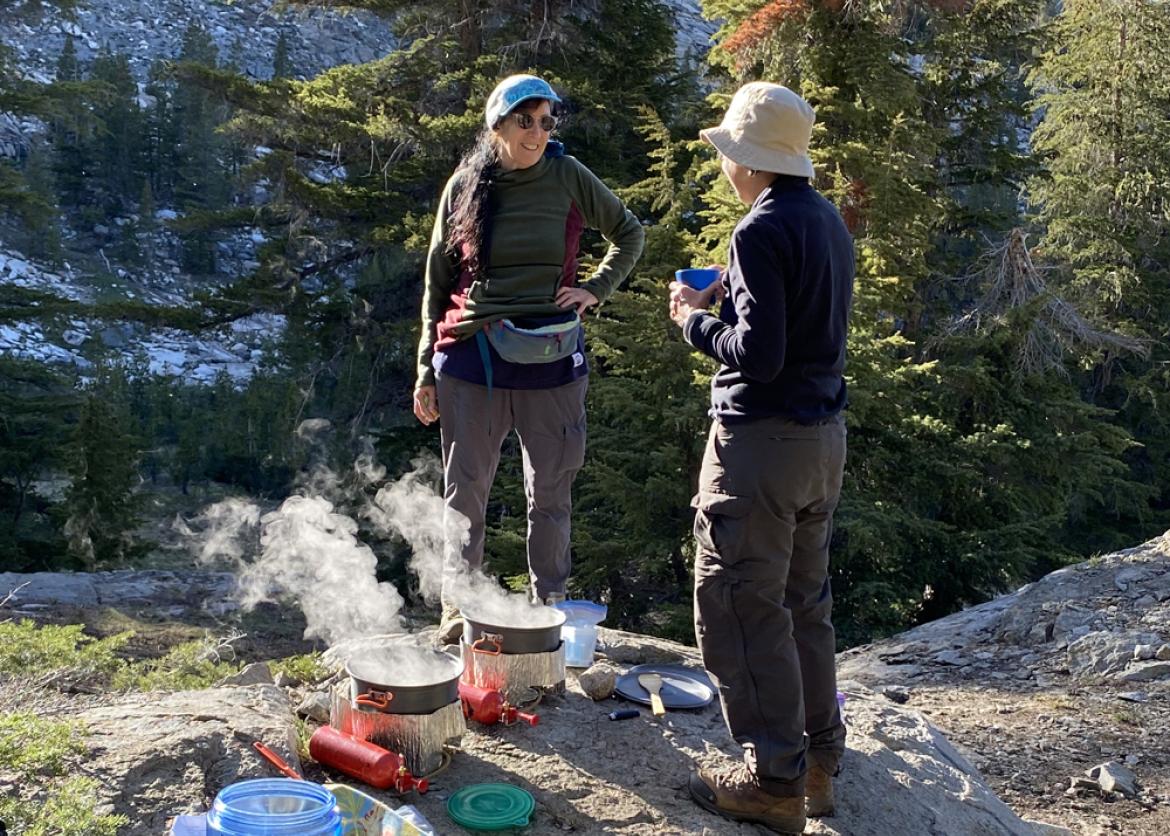 Two women standing on a large rock with their portable stove and pots as they cook and chat