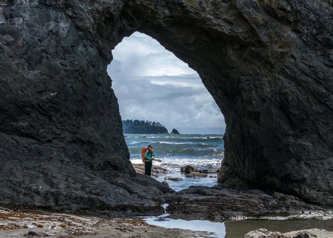 A person seen through a naturally forming stone arch on the beach.