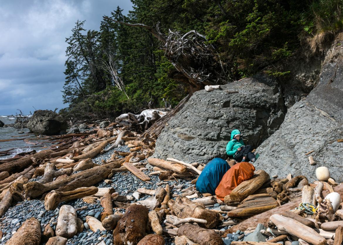 A person wearing a rain slicker resting and reading on a driftwood covered beach.