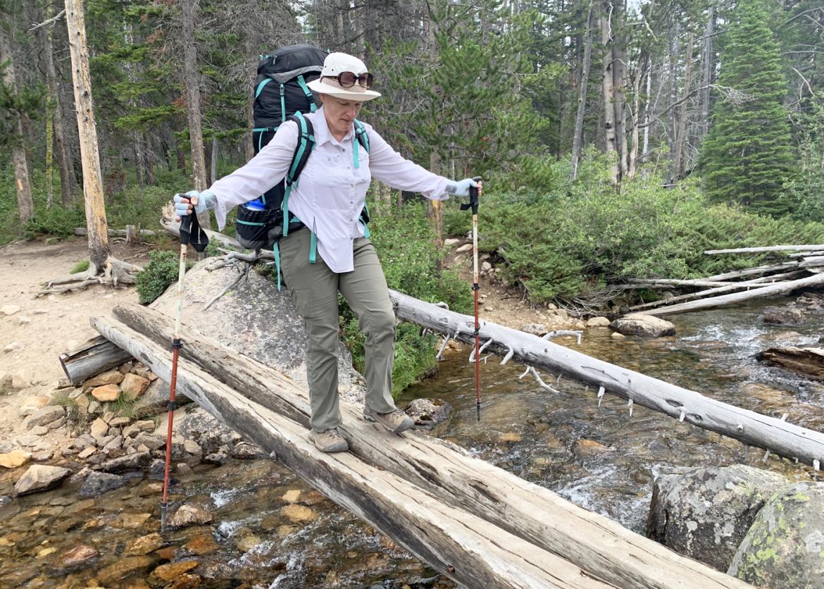 A woman crosses a creek. She walks on a log, balancing herself with hiking poles.