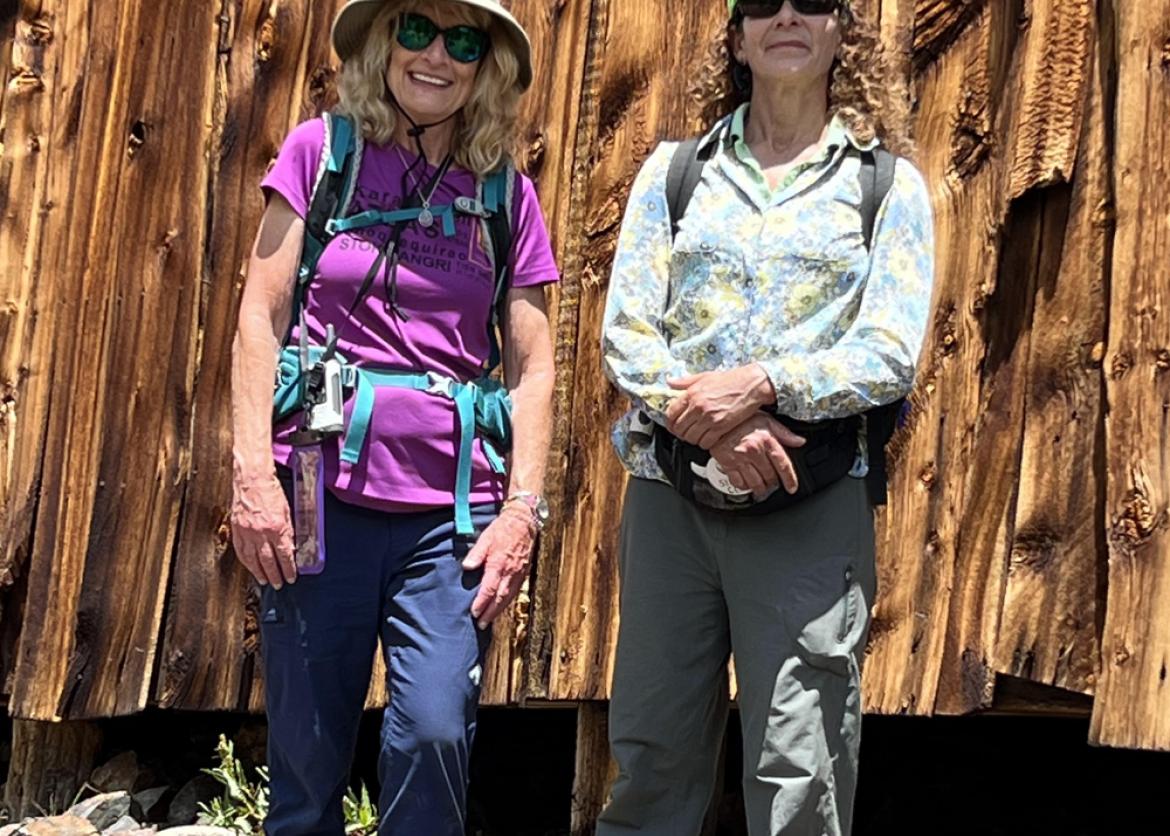 Two women in outdoor wear stand in front of a wooden wall.