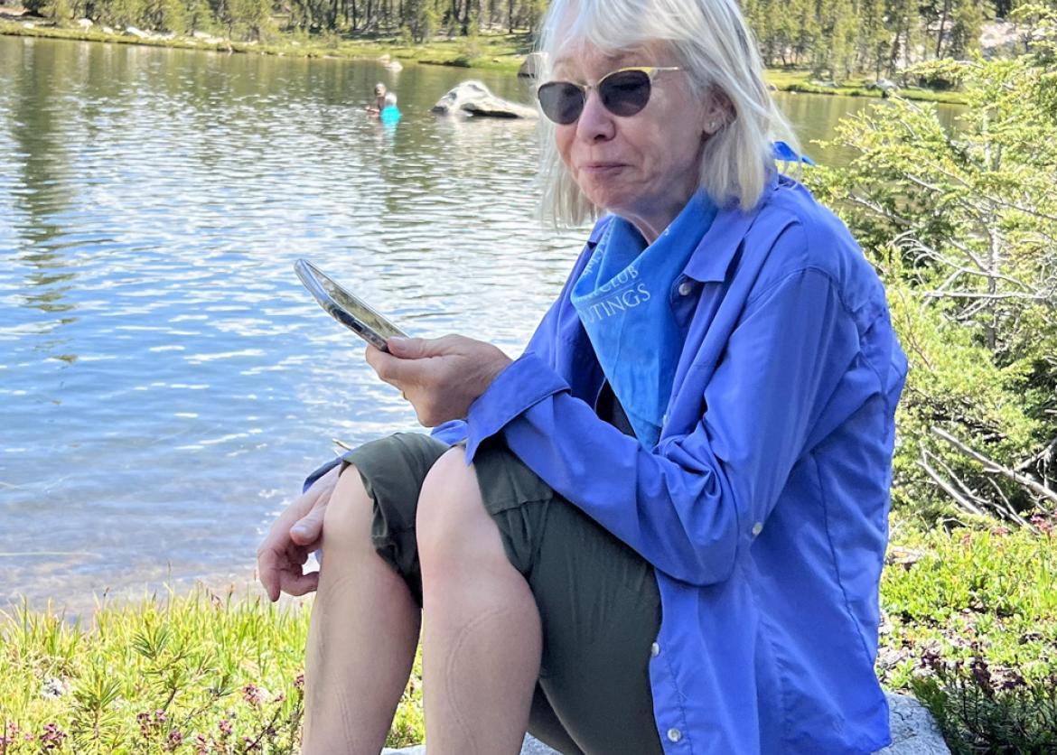 A woman sitting barefoot on a rock by a lake. She checks her phone. To the side is a partially eaten apple.
