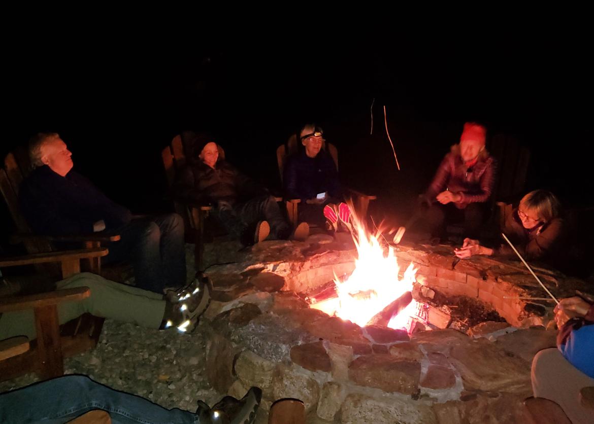 A group surrounding the campfire smiling and chatting in the dark.