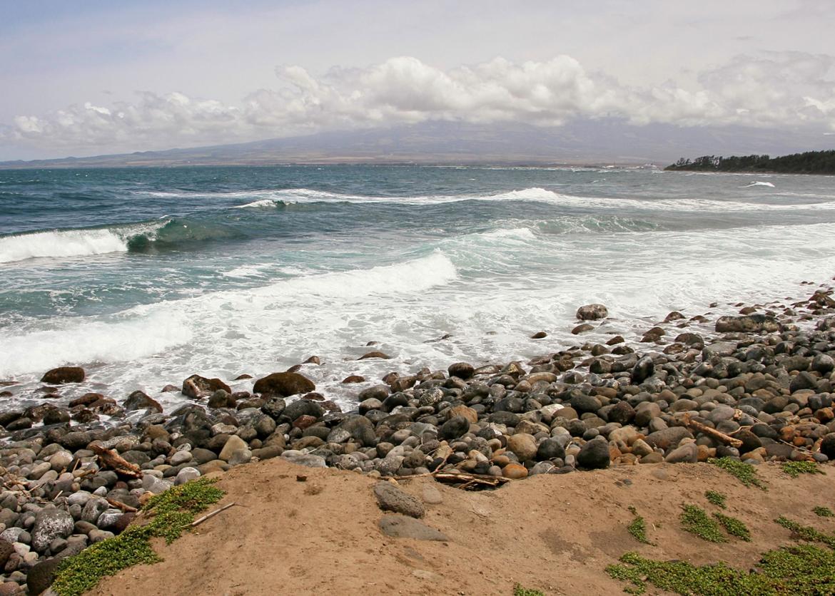 A view looking out towards the ocean. Large round rocks in the foreground, mild waves crashing down, and a mountain way off in the distance covered in white puffy clouds.