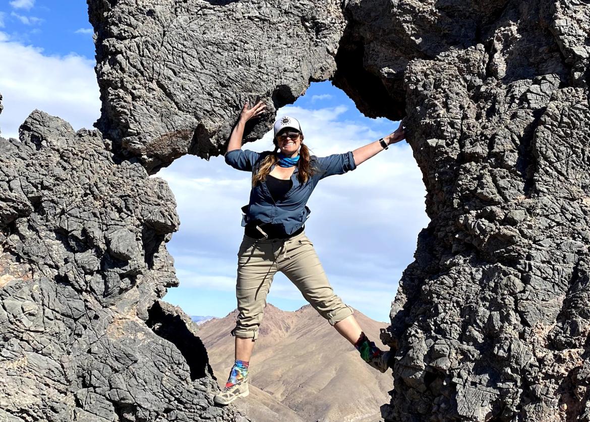 A smiling woman in outdoor gear stands in a hole within a rock formation.