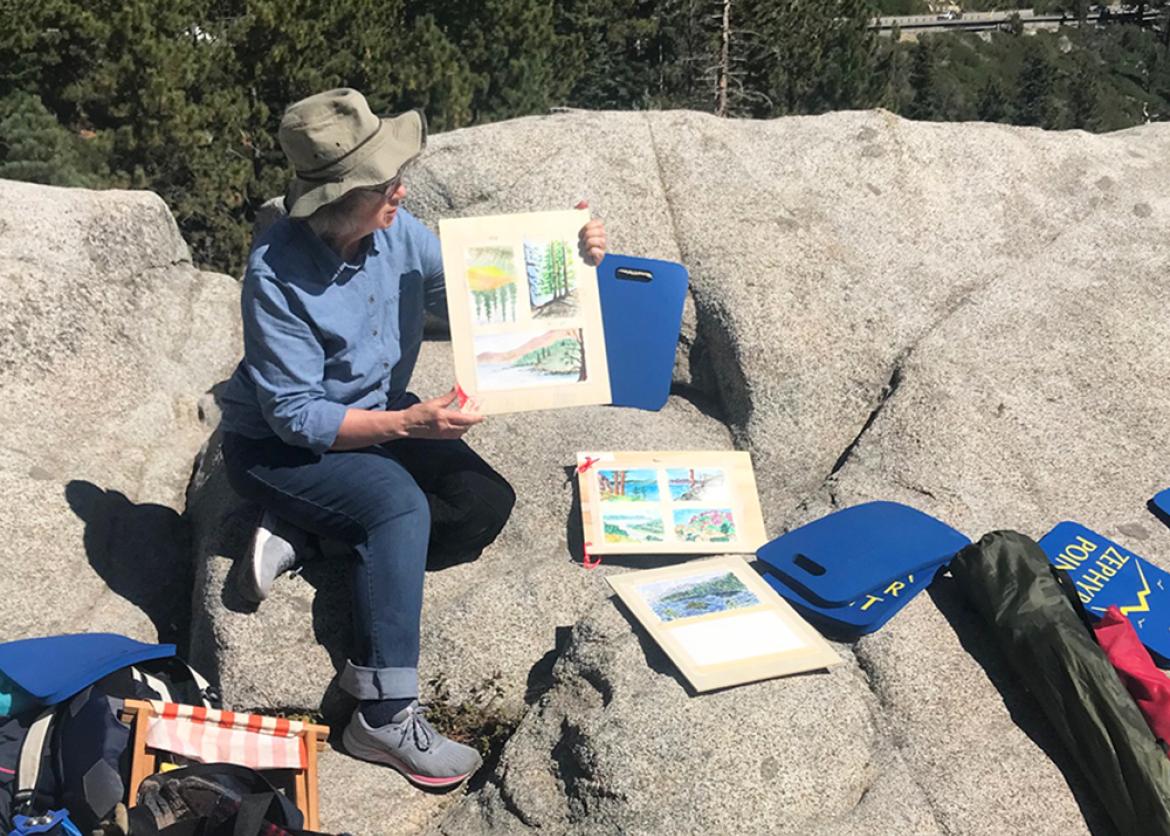 Woman in the outdoors displaying artwork and surrounded by art supplies.