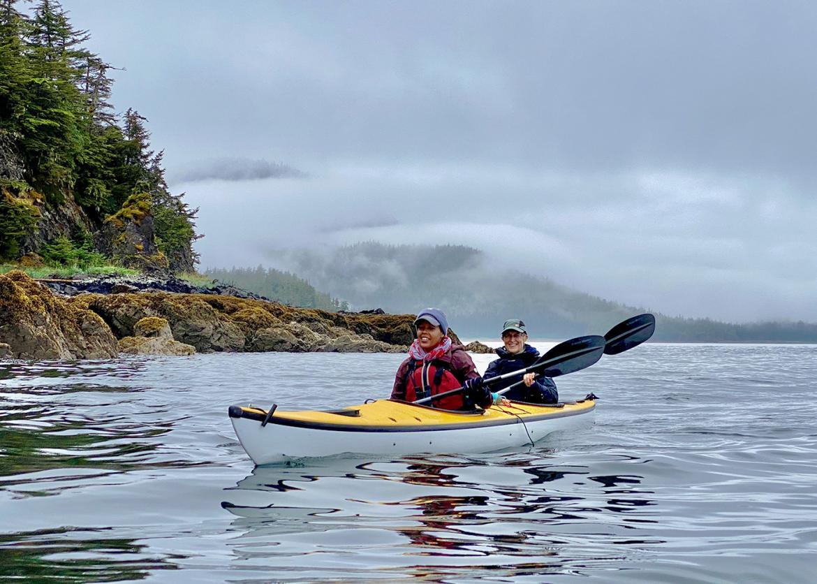 Two people in a double kayak float next to a forested coast on a foggy day.