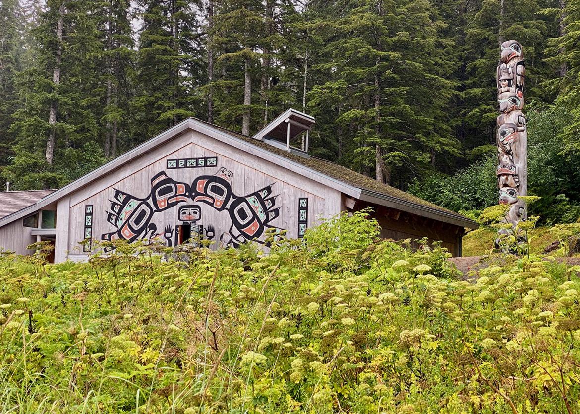 A totem pole and a house decorated with traditional painted artwork sits within a forest.