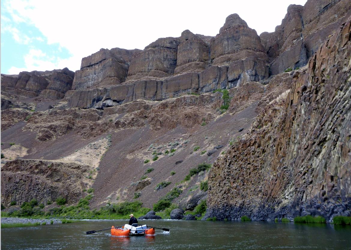 A person on a boat stocked with coolers rows in front of a tall rocky slope.