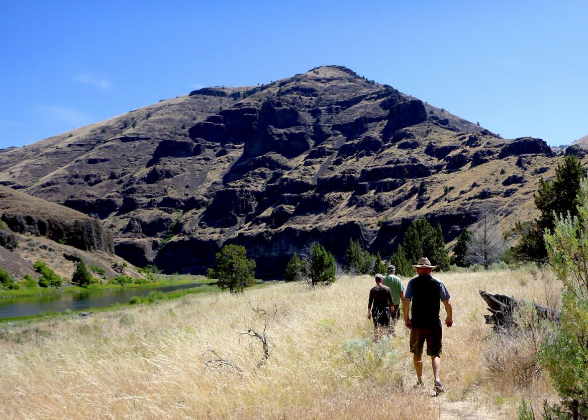 Three hikers walk on a trail through dry grass and towards a large hiill.
