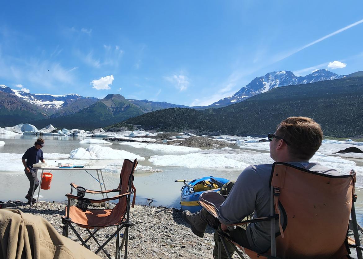 Trip participants relaxing at camp in front of icy lake, with mountains in the background