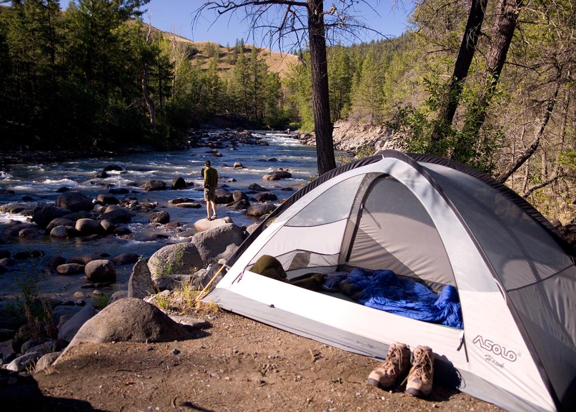 Camping tent in a forest in the foreground with person standing by river in the background