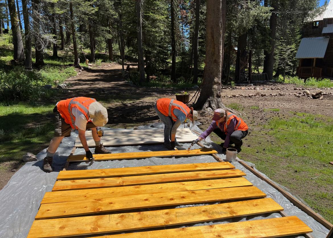 Three participants staining long wood boards, near the lodge in the forest