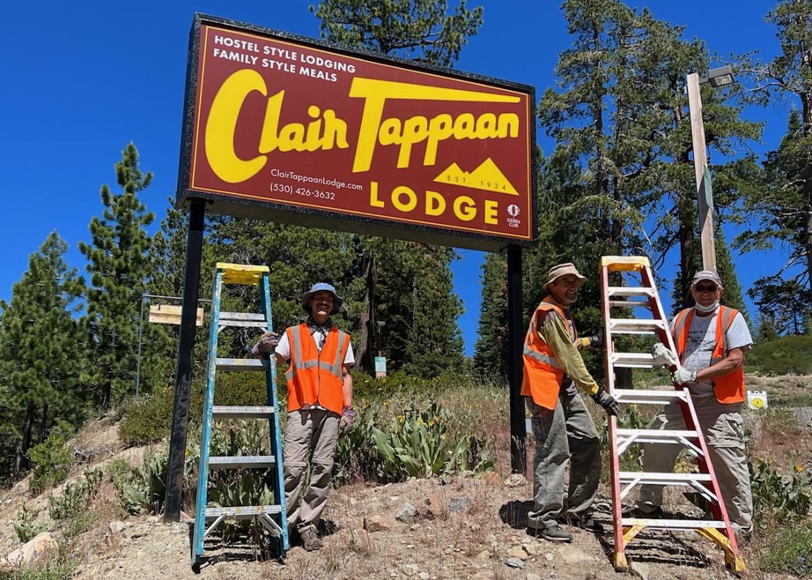 A group of 3 participants standing with ladders while working on a hotel sign, pine trees in the background