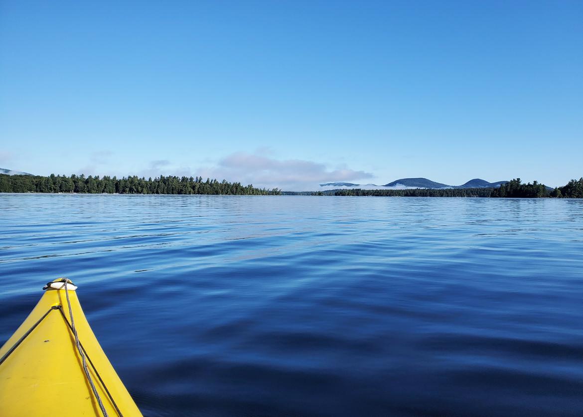 Tip of a yellow kayak on a calm blue lake in the Adirondacks of New York.