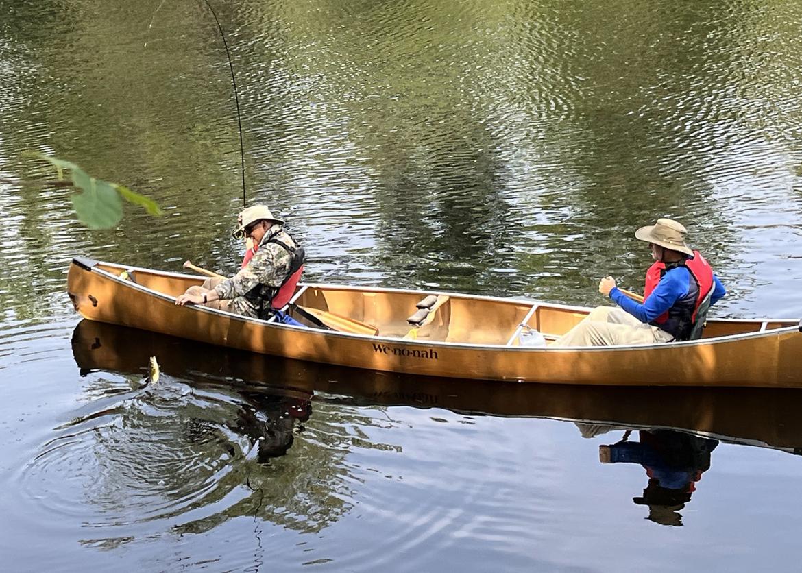 Photo: Two people in a canoe on the water, one man holds a fishing pole and is reeling in a fish