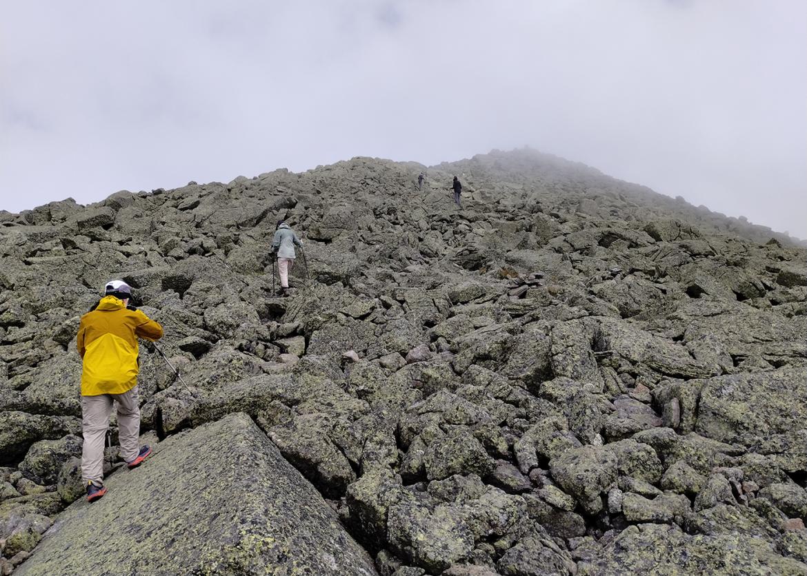 Hiker in yellow jacket climbing up a rock field towards the summit of a mountain