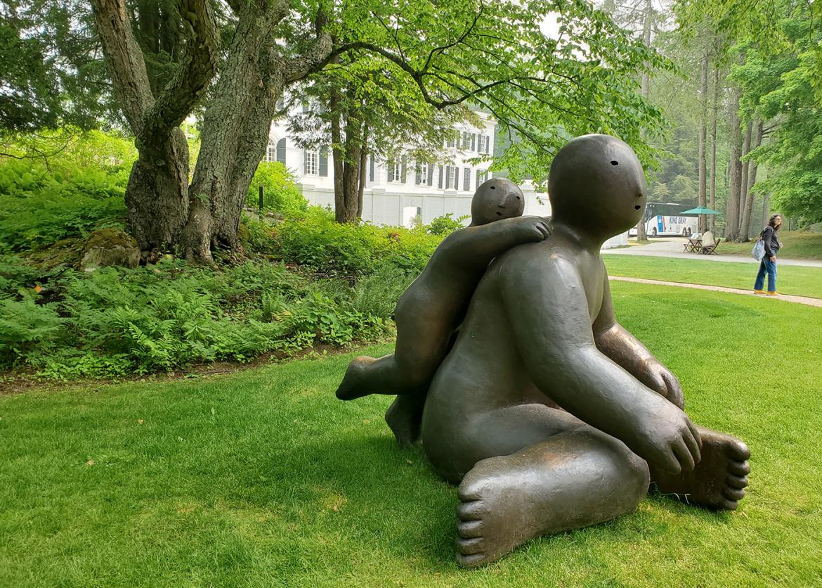 Large modern sculpture of an adult human figure with child on back, in a garden surrounded by grass and trees.