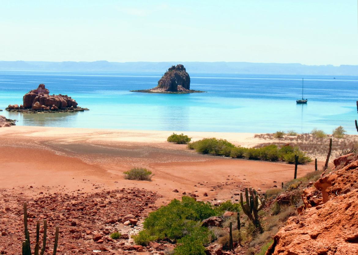 A desert shoreline.  Two rocky islands and a sail boat jutt out of the water.