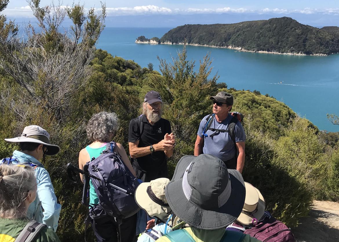 A group of hikers converse. The background view is of pristine waters and a forest covered island.