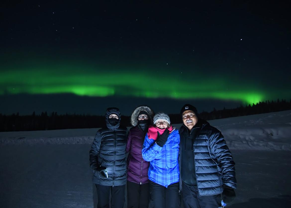 Four smiling people posing in front of a view of the Aurora Borealis.