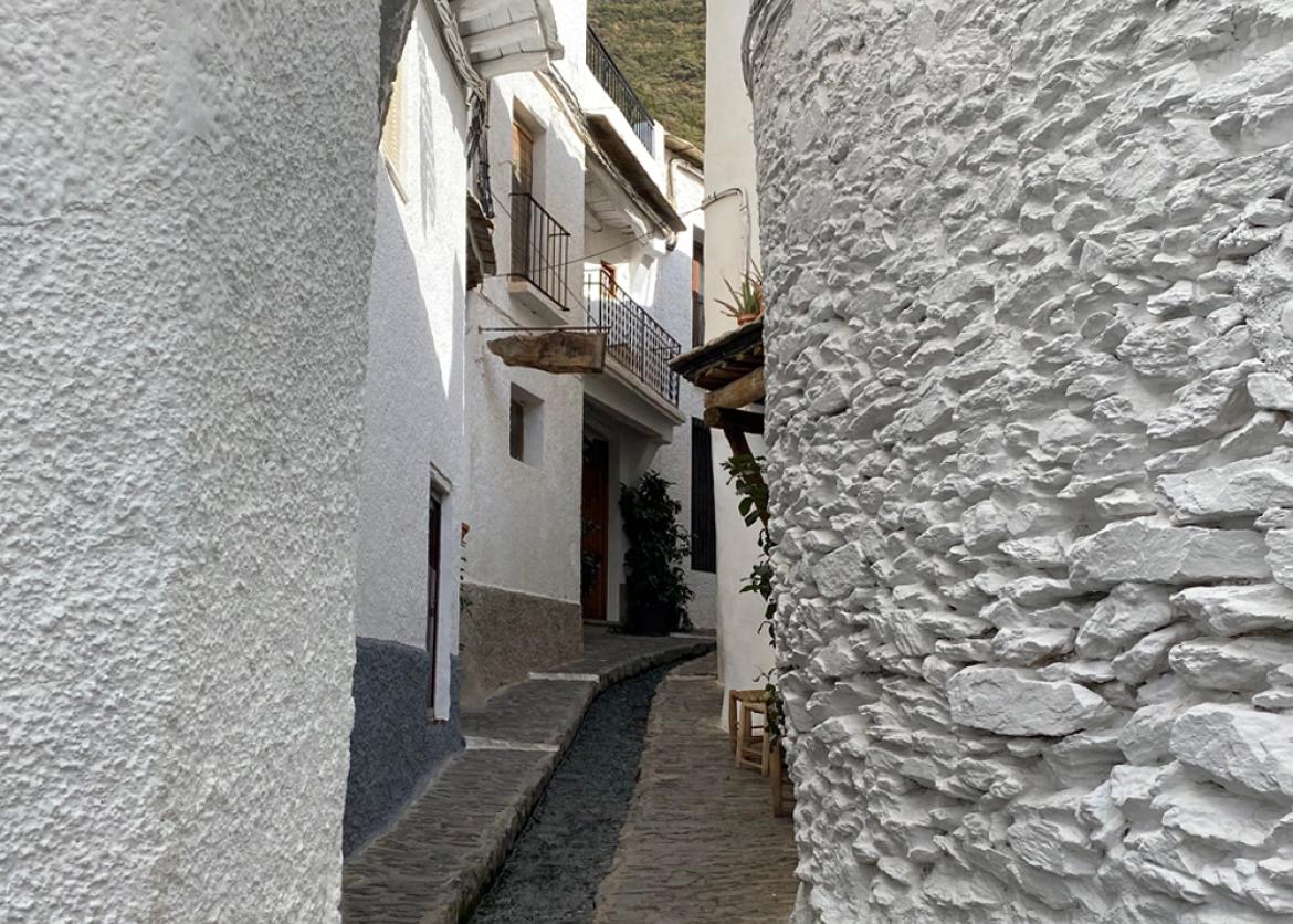 A stone paved alleway with a gutter winding between white buildings.  A brightly colored woven decoration hangs above.