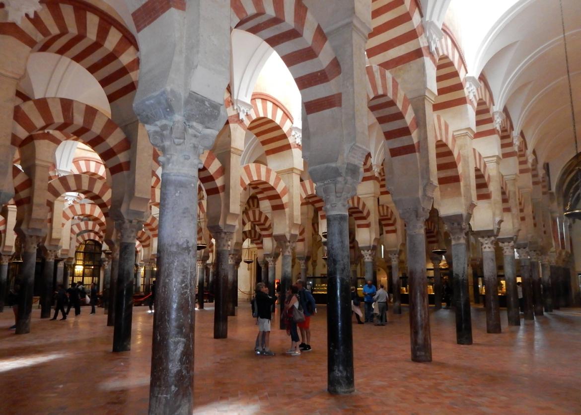 A room full of columns connected by ceiling arches.  The arches are patterned with red and white stripes.  People stand in clusters between the columns.