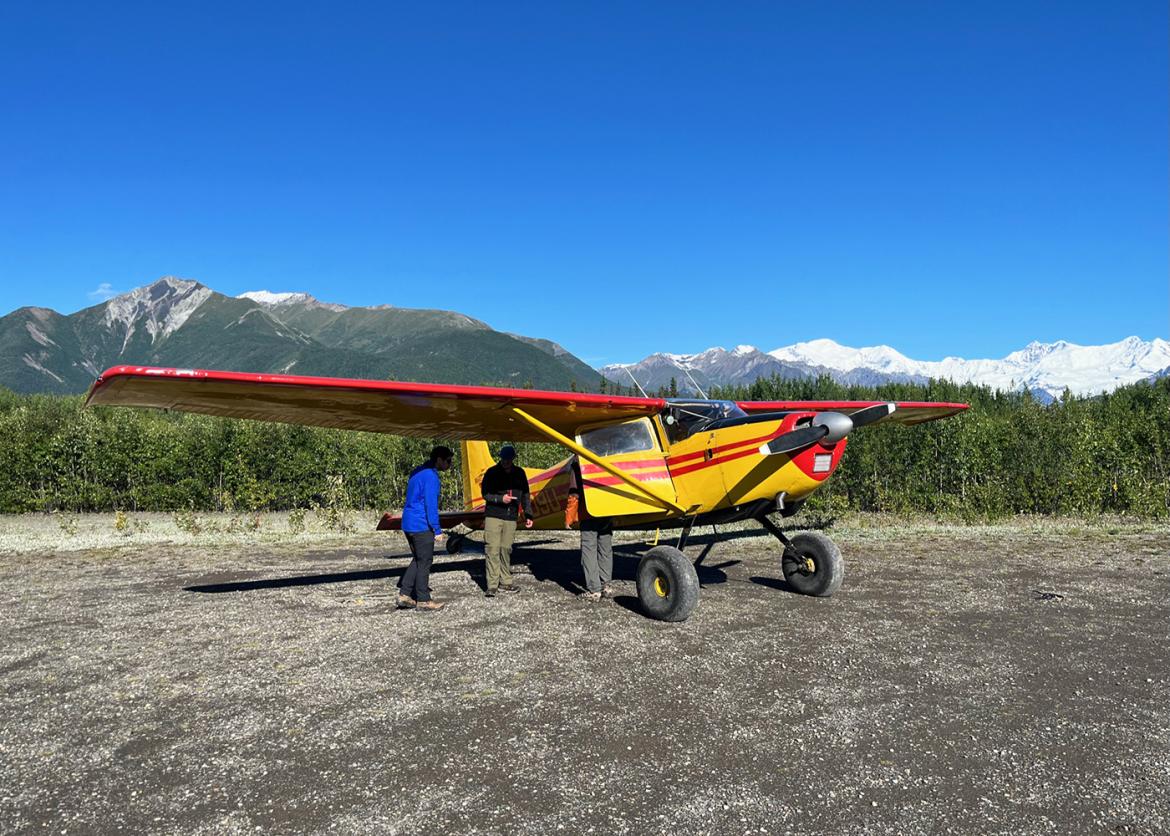 Three people in front of small yellow bush plane with snowcapped mountains in background