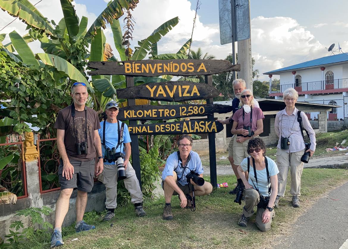 A group of people all wearing binoculars and cameras stand in front of a sign which reads "Bíenvenídos a yaviza, kilometro 12,580, final desde Alaska."