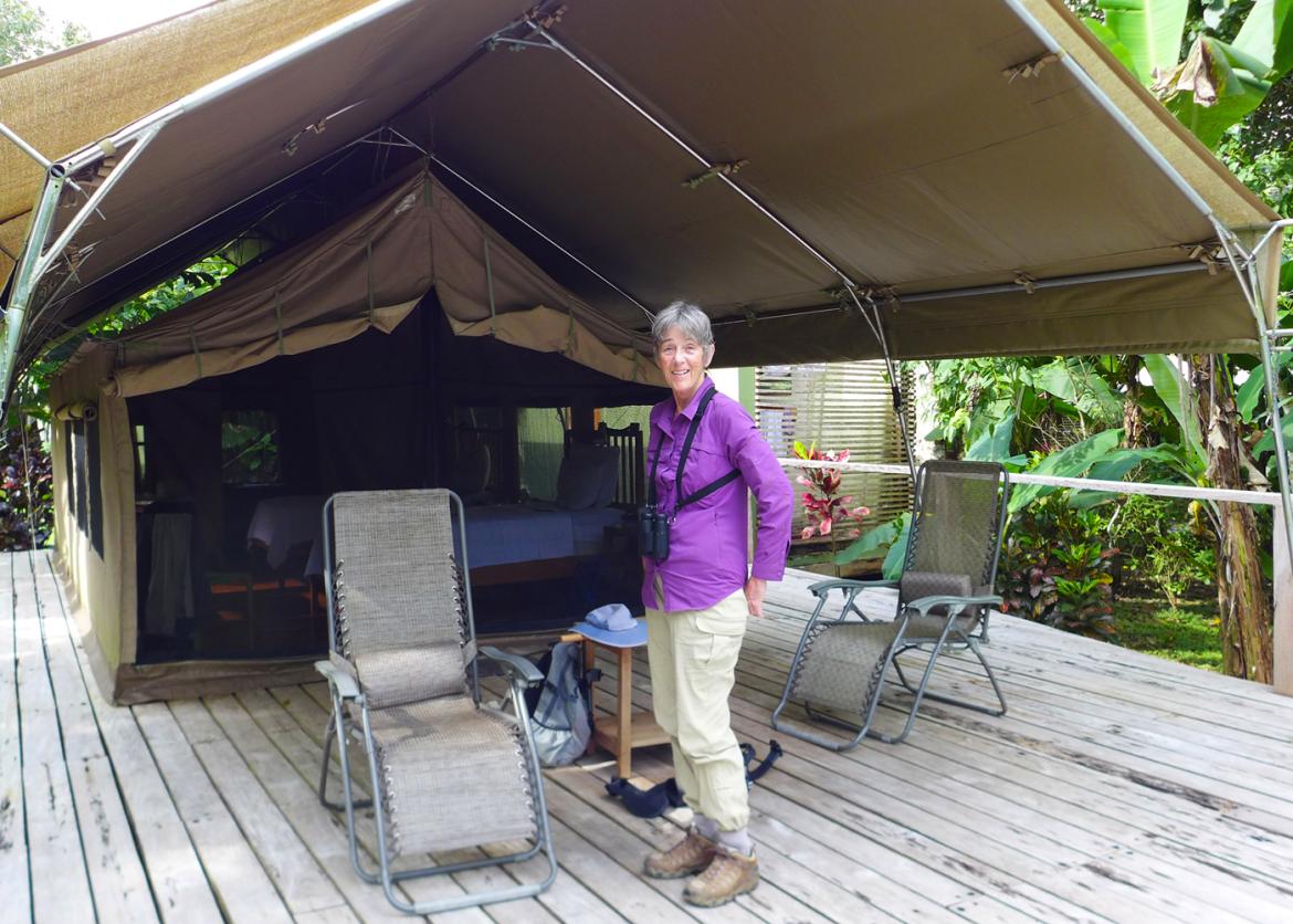 An individual stands on a wooden platform in front of a large tent.  Two single beds are in the tent.  A canopy covers the tent as well as two deck chairs.