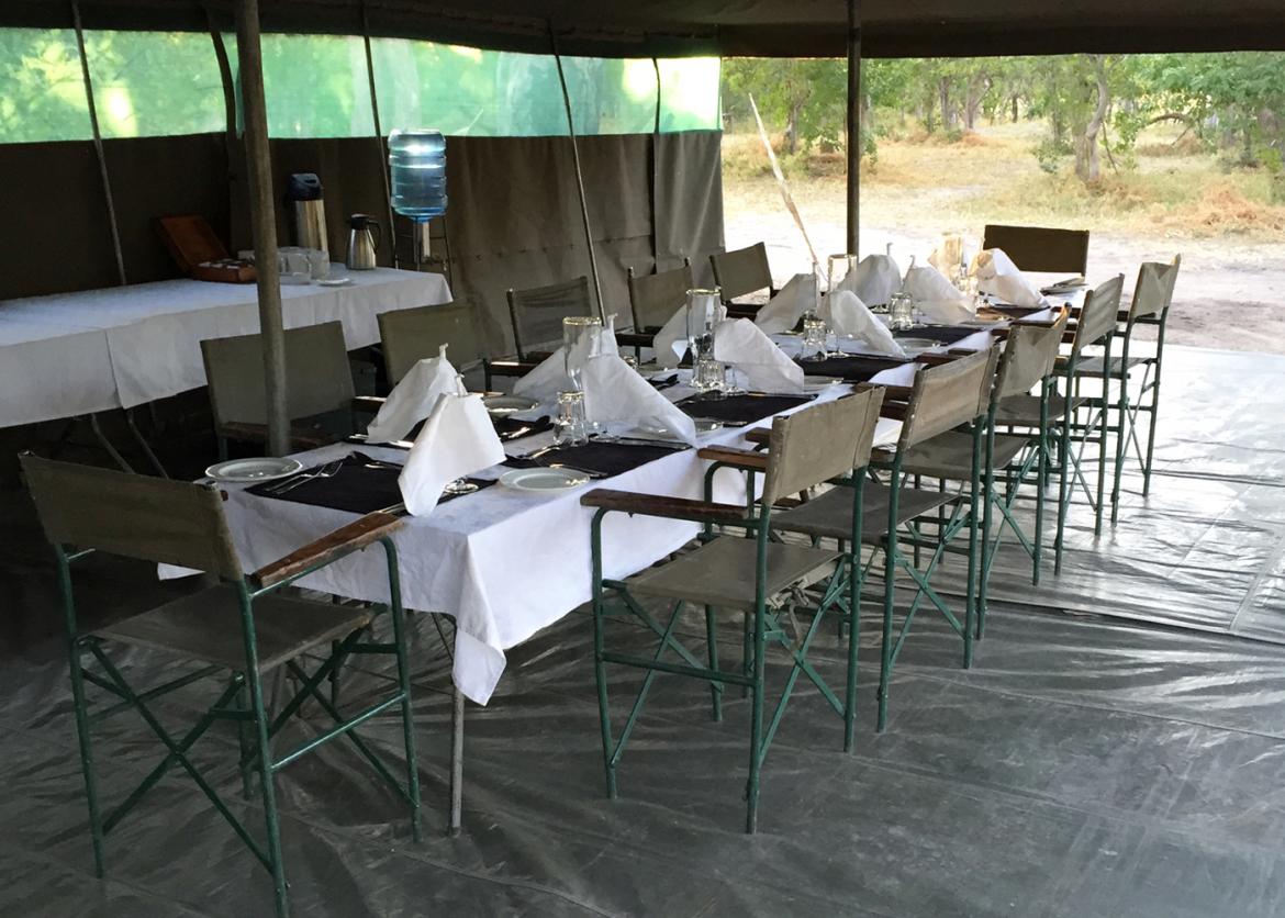 A long table holding table settings and wine glasses and surrounded by folding chairs.  The table rests on a tarp and underneath a large tent.
