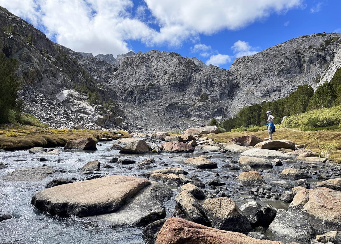 Single hiker standing on rocks in a wide river, surrounded by beautiful mountain scenery